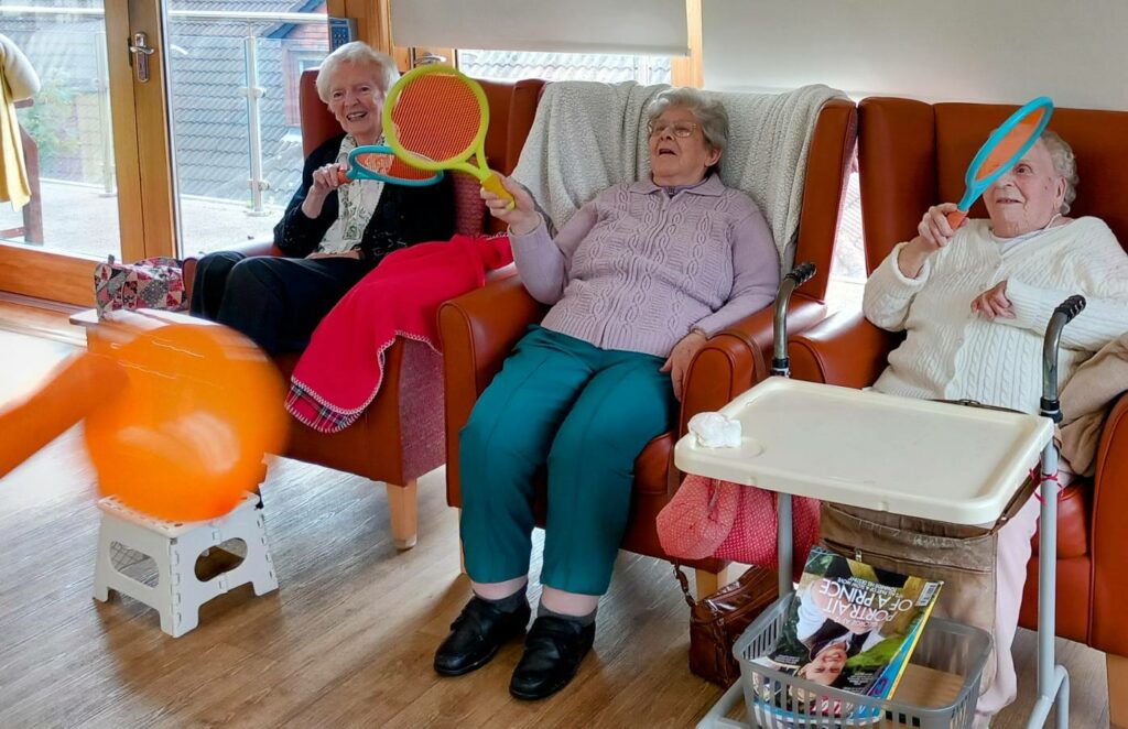 Three elderly women sit in armchairs indoors, holding colourful rackets and playing with a large orange ball. A walking frame and a tray with magazines are visible nearby.