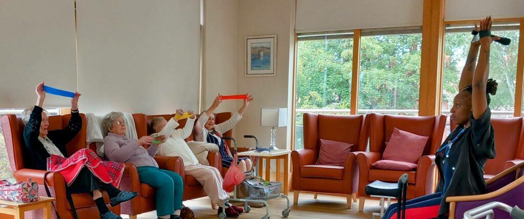 Four elderly people seated on armchairs hold coloured exercise bands above their heads, following instructions from a facilitator standing in front of them in a bright room.