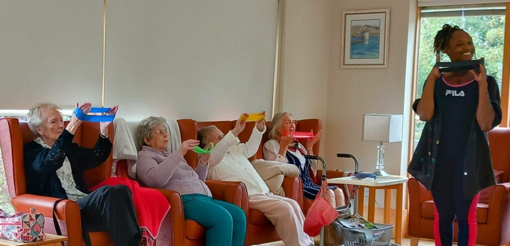 Five elderly women seated in armchairs take part in a group exercise, holding coloured bands, while an instructor stands before them in a well-lit room.