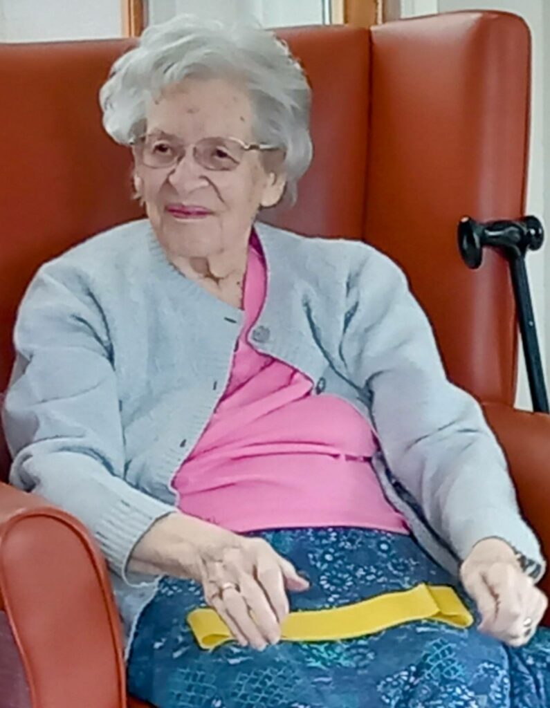 An elderly woman with grey hair and glasses sits in a red chair, wearing a pink top, grey cardigan, and blue skirt, with a yellow resistance band and a black walking stick next to her.
