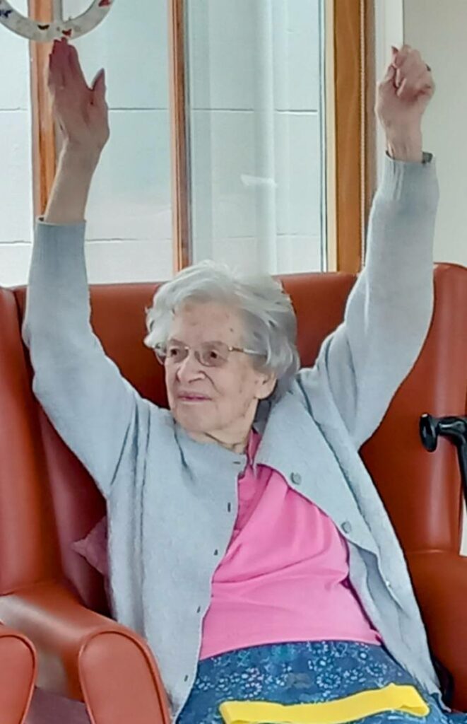 An elderly woman with grey hair and spectacles sits in a red chair, lifting both arms above her head. She wears a pink top and grey cardigan. A walking frame is visible beside her.