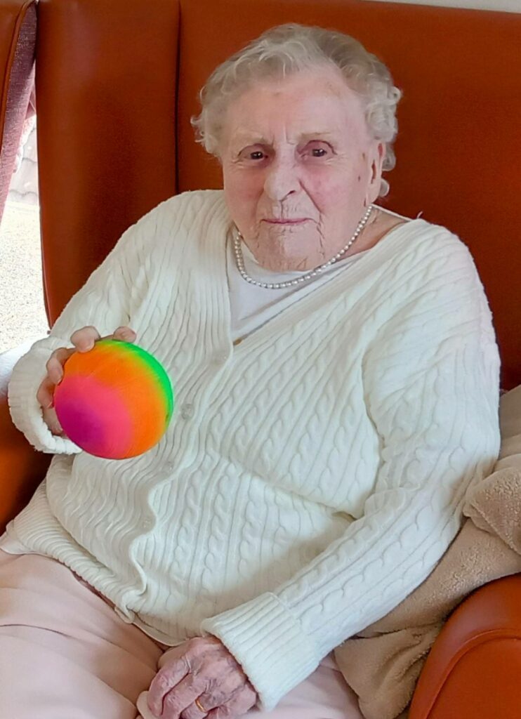 Elderly woman seated on a brown chair, wearing a white jumper and holding a colourful, rainbow-patterned ball.