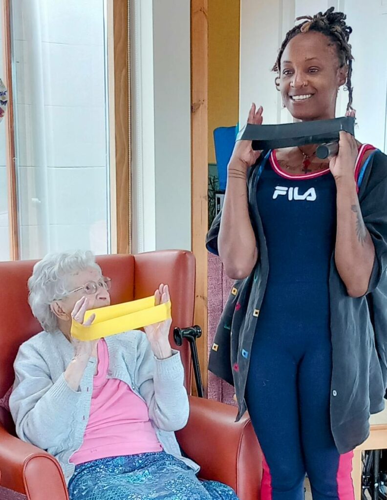 An elderly woman seated in a chair and a younger woman standing beside her each hold a rectangular object to their mouths and smile in a brightly lit room.