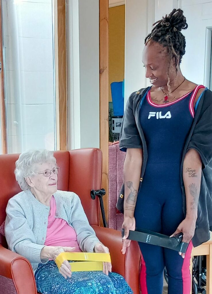 An older woman sits in a chair holding a yellow box, while a younger woman in sportswear stands beside her, smiling and holding a black box.
