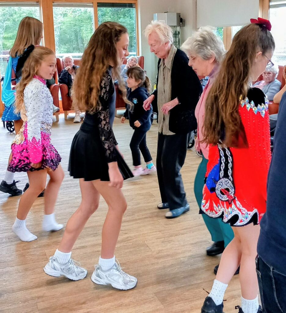 Several young girls in traditional Irish dancing costumes and older women are dancing together indoors at Abbeyfield, while people seated in the background look on.