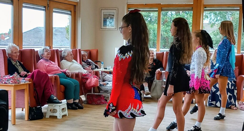A group of girls in colourful Irish dancing costumes perform for a seated audience of elderly women at Abbeyfield, in a bright room with large windows.