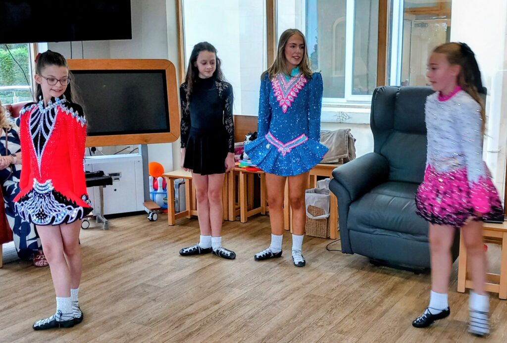 Four girls in colourful Irish dancing costumes and soft shoes stand in an Abbeyfield room with wooden flooring, a television, chairs, and a window in the background.
