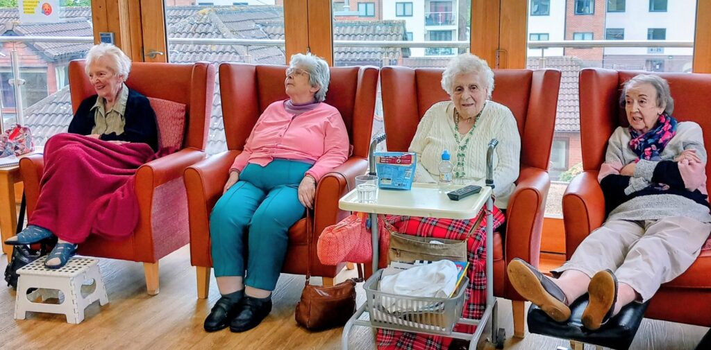 Four elderly women sit in armchairs in the Abbeyfield communal lounge, one draped in a red blanket and another with a walking frame; a table with bottled water is before them as they chat about Irish dancing.