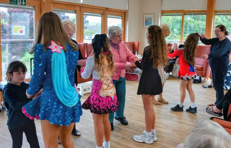 Young girls in colourful Irish dance costumes and elderly women are enjoying Irish dancing together in the bright Abbeyfield community room, with large windows and seated onlookers cheering them on.