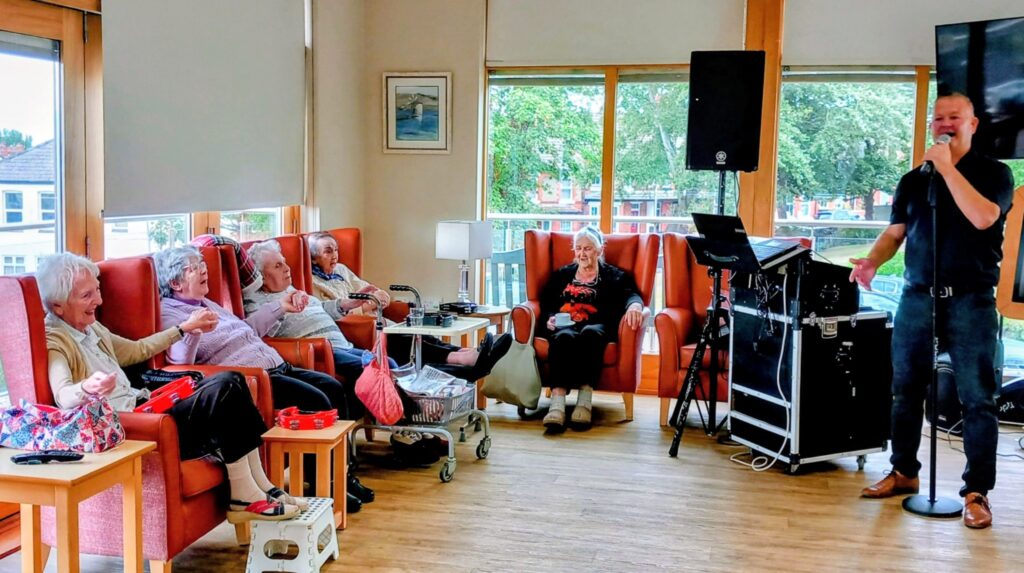 A group of elderly people sit in armchairs in a bright room, watching a man sing into a microphone beside audio equipment.
