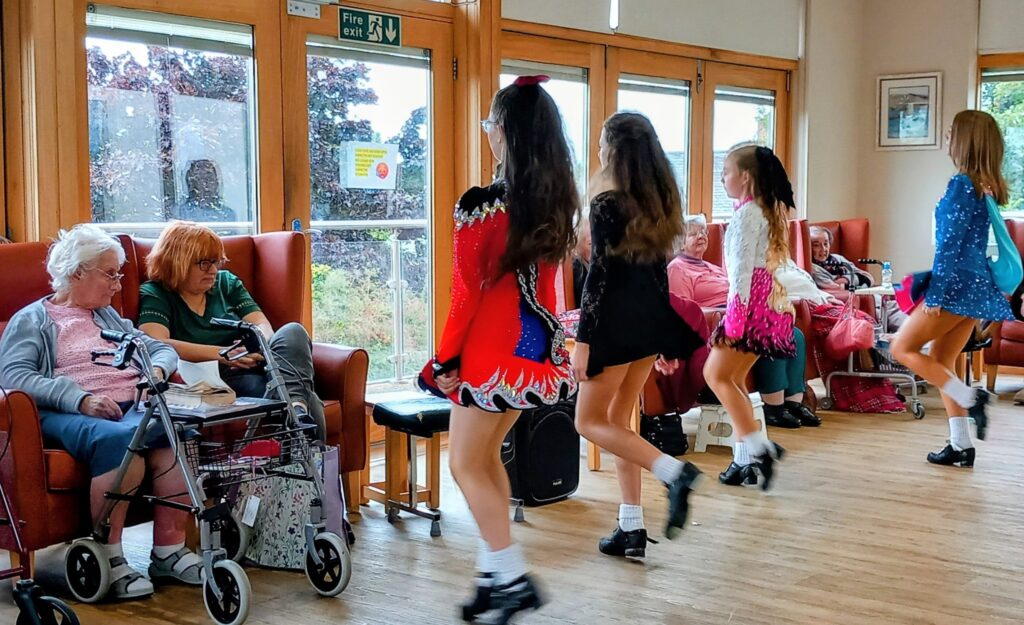 Four girls in colourful costumes perform Irish dancing in an Abbeyfield care home lounge, bringing a joyful afternoon to elderly residents who watch from their chairs.