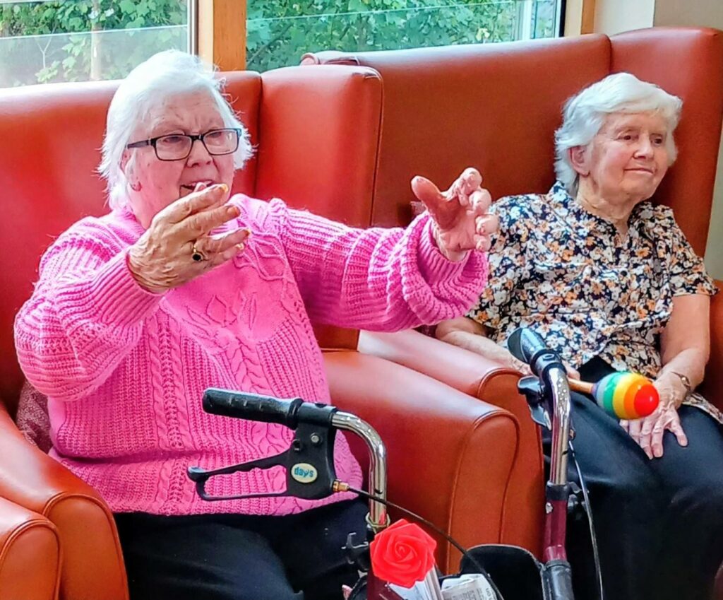 Two elderly women sit on orange chairs; one gestures with her hands while the other sits calmly beside a walking frame topped with a toy and a red rose.