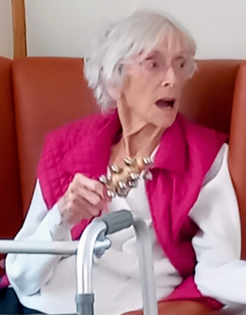 Elderly woman with white hair and spectacles, wearing a pink waistcoat, sits in a chair holding a hand bell, with a walking frame in front of her.