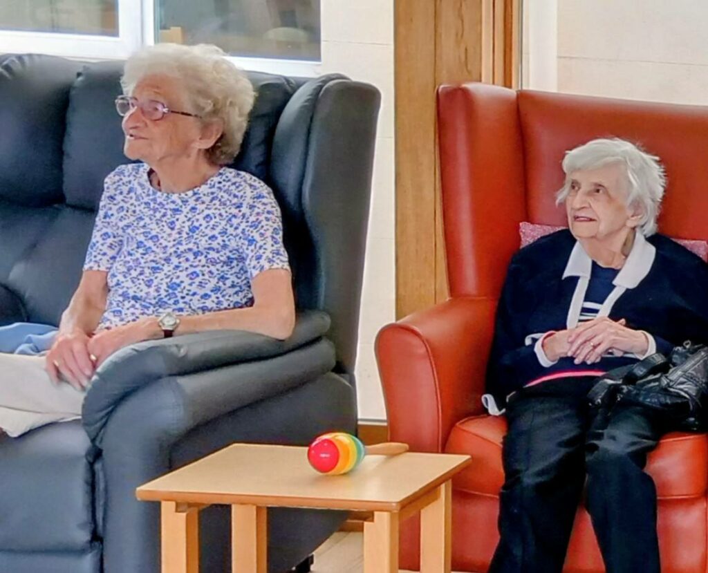 Two elderly women sit in armchairs, one grey and one red, with a small table and a colourful toy in front of them. Both are looking slightly to the left.