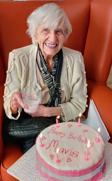 An elderly woman sits in an orange chair, smiling and holding a glass, with a birthday cake in front of her that reads Happy Birthday Mavis and has six lit candles.