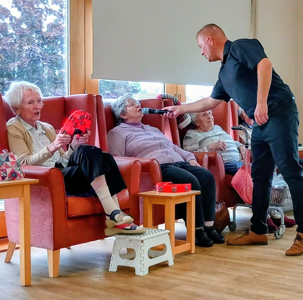 A man holds a microphone towards an elderly woman seated in a care home lounge, while two other older women sit nearby on armchairs.