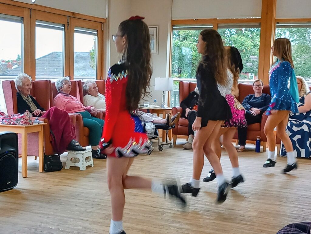 Four young dancers perform Irish dancing in colourful costumes for a group of seated elderly women at Abbeyfield, in a bright and spacious room with large windows.