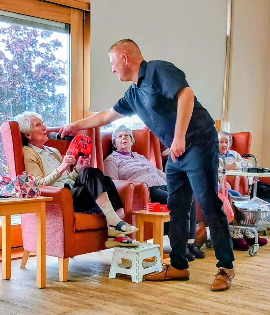 A man holds a microphone for an elderly woman seated in a chair, with two other elderly women sitting nearby in a communal room.
