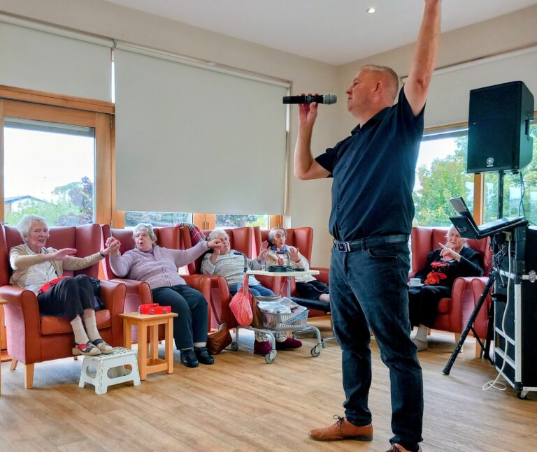 A man sings into a microphone while elderly people seated on chairs watch and raise their arms in a brightly lit room.