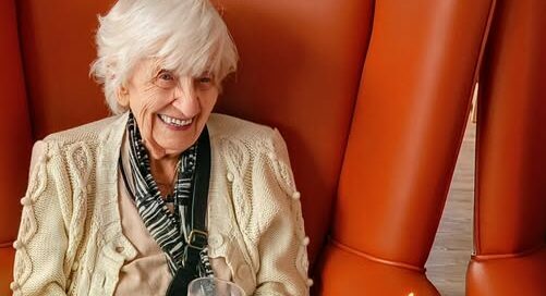 Elderly woman seated on an orange chair, smiling and holding a glass, with a birthday cake topped with lit candles in front of her.