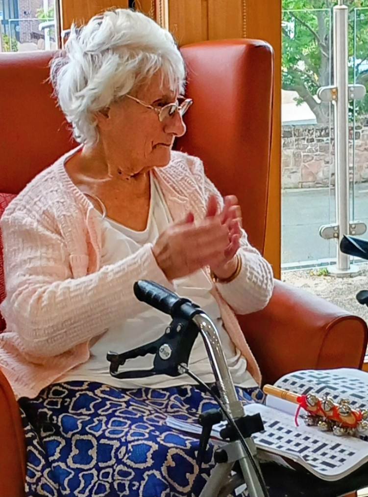 Elderly woman seated in an armchair, applauding with her hands, a walking frame beside her and a bingo card on her lap.
