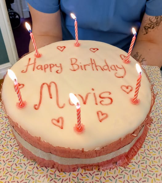 A round birthday cake with white icing, pink writing reading Happy Birthday Mavis, six lit candles, and small heart decorations—ideal for marking Mavis’s special day of celebration.