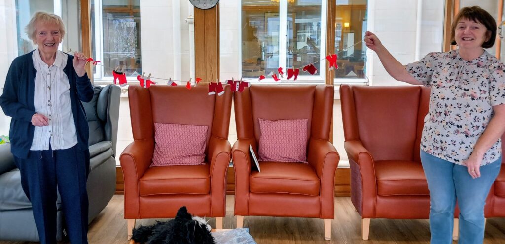 Two women stand smiling, each holding one end of a string adorned with small red and white decorations, celebrating Christmas Shoeboxes, in front of three empty armchairs in a bright Wirral room.