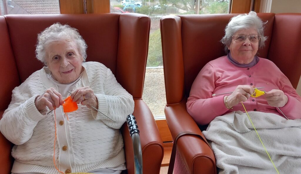 Two elderly women sit side by side in armchairs, knitting orange and yellow wool for Christmas Shoeboxes, in a bright room with large windows, sharing warmth with the world.