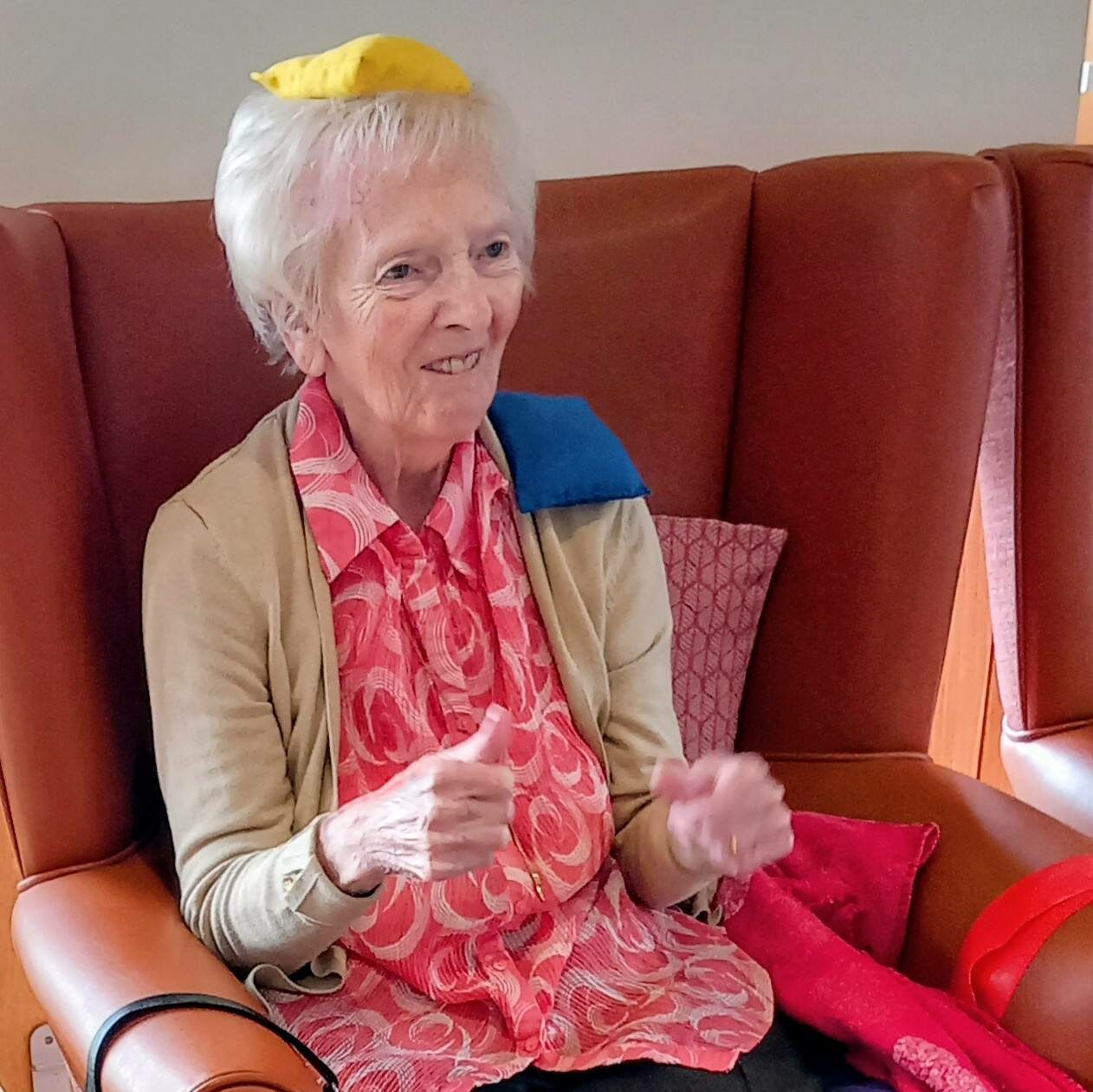A strong older woman sits in an armchair with coloured beanbags balanced on her head, shoulder, lap, and foot. She smiles warmly and is dressed in casual clothes.
