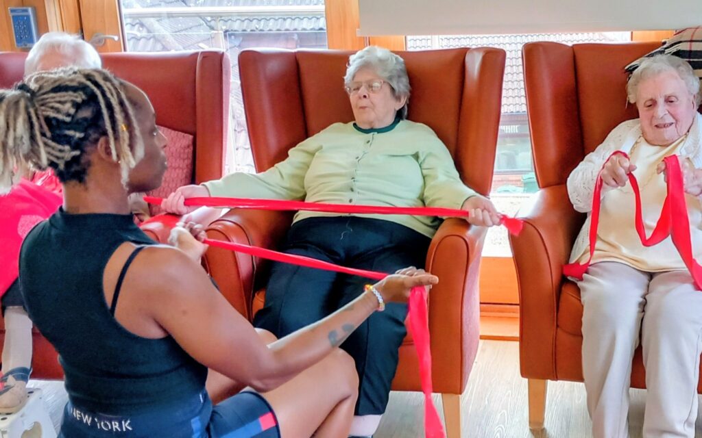 A group of smiling elderly women seated on chairs exercise with red resistance bands, guided by an instructor in a well-lit room at Abbeyfield.