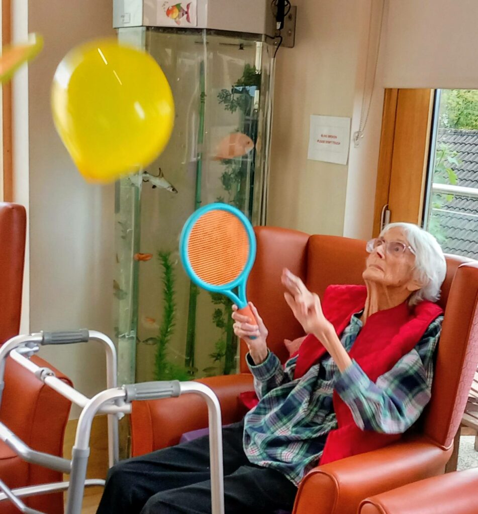 A smiling elderly woman at Abbeyfield shows her strength as she uses a blue bat to strike a yellow balloon, with her walking frame close by and a fish tank in the background.