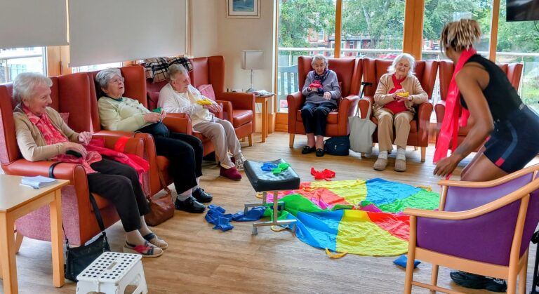 Six elderly women sit in armchairs in a bright Abbeyfield room, smiling as a facilitator leads a keep fit activity with colourful fabric and a parachute on the floor.