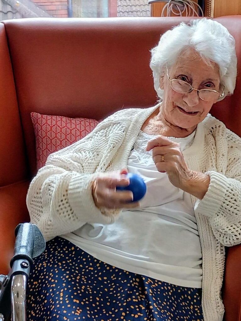 Elderly woman with white hair and glasses sits on a red sofa at Abbeyfield, smiling and holding a small blue ball in her hand. She looks robust in her white jumper and patterned skirt.
