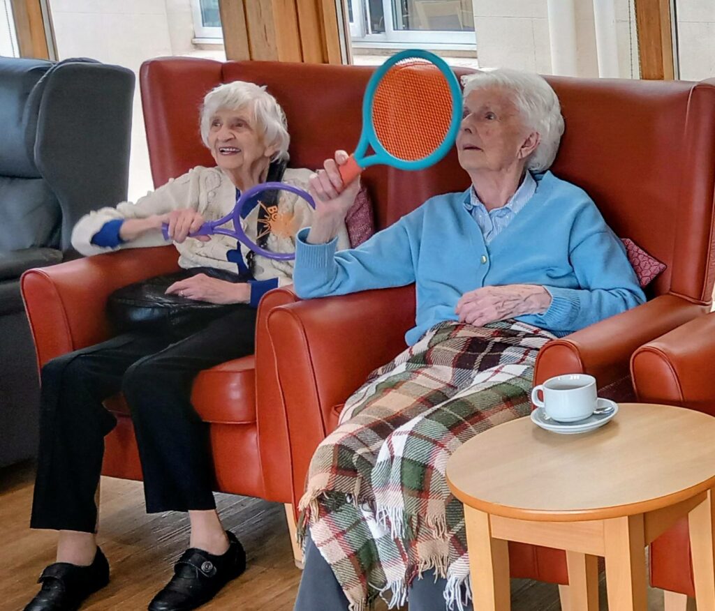 Two elderly women sit on armchairs indoors, each holding a plastic racket as if mid-game. At Abbeyfield, one is smiling with a blanket across her lap; a cup and saucer sit on a small table nearby.