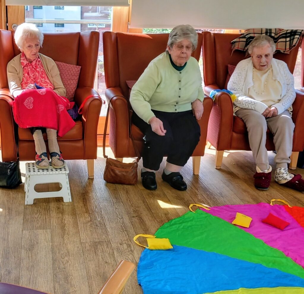 Three smiling elderly women sit in armchairs at Abbeyfield, facing a colourful parachute mat with bean bags on the floor, enjoying activities to help them stay strong.