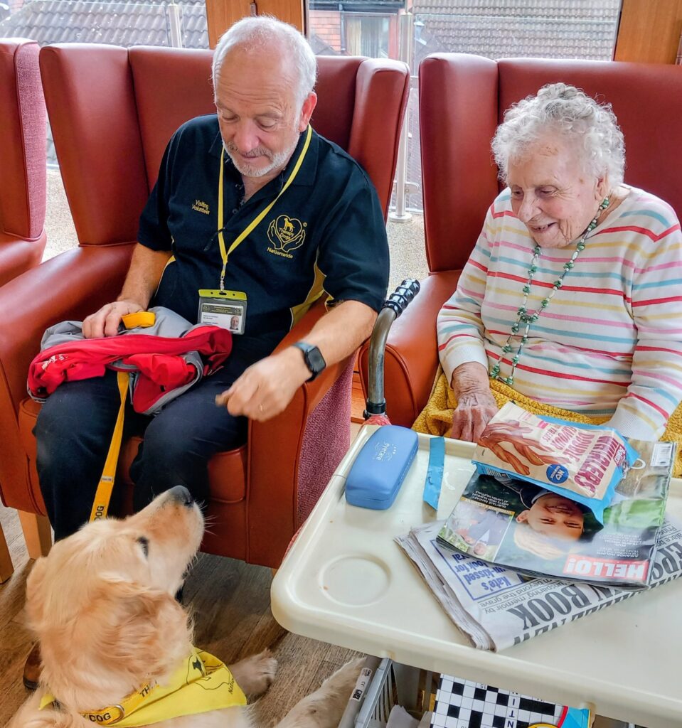 An elderly man and woman sit in armchairs, engaging with a golden retriever during a cosy Pawsitive Afternoon. Newspapers, snacks, and personal belongings are on the table in front of them.