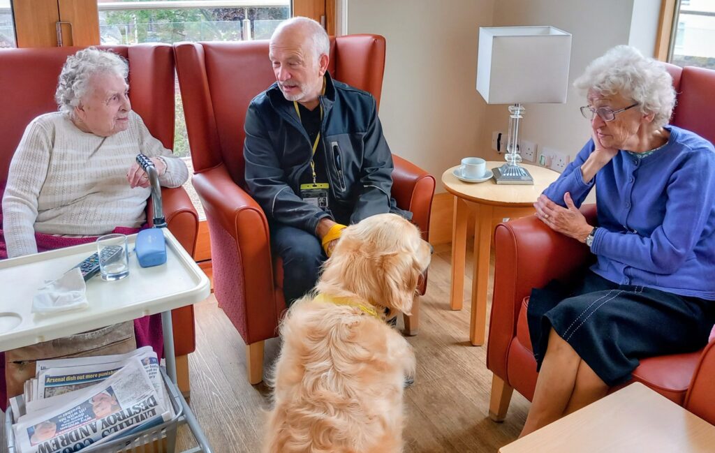 Three elderly people sit in armchairs, chatting with a man while a golden retriever faces them at the Pawsitive Afternoon pet event. In the foreground, a tray holds newspapers, water, and a blood pressure monitor.