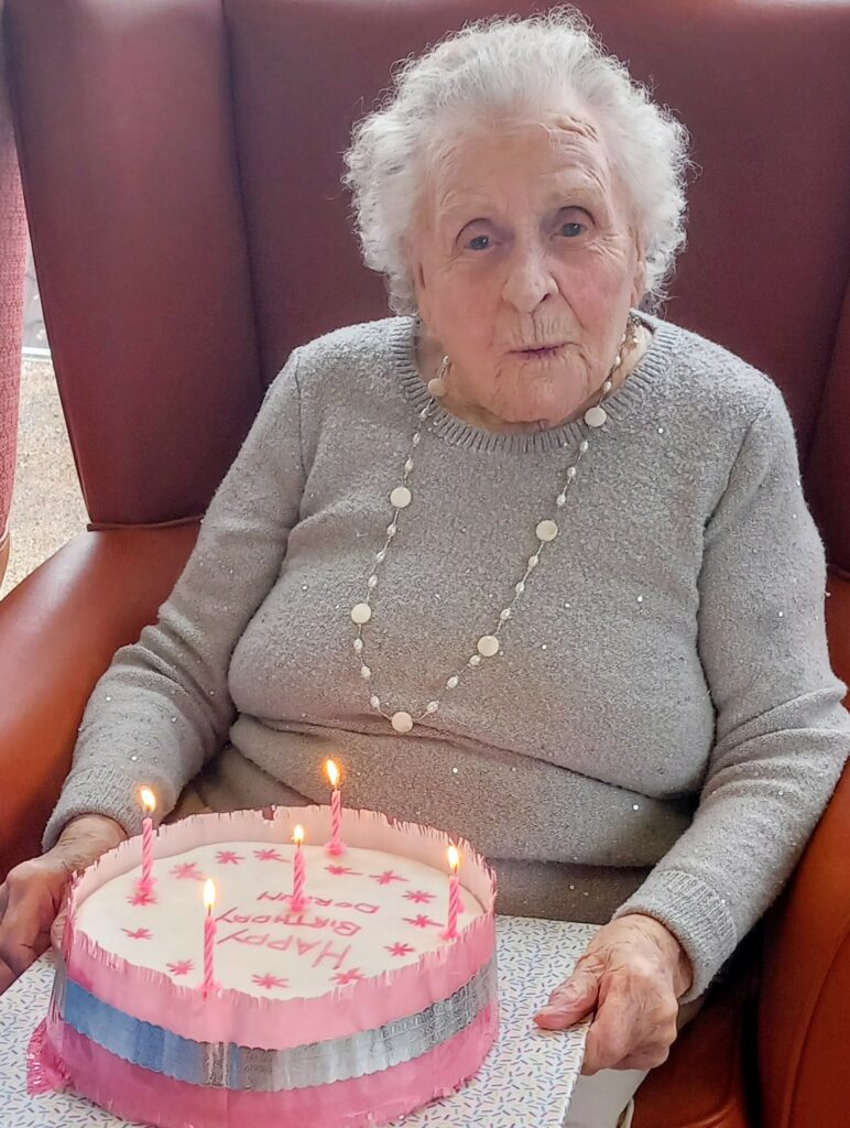 Elderly woman seated in a chair holding a pink birthday cake with lit candles and 'Happy Birthday' written on it.