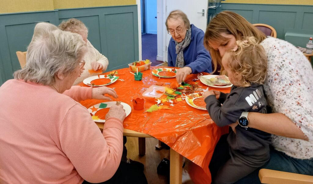 Several elderly women, a young woman, and a child sit around a table draped with an orange cloth, making colourful paper shapes and enjoying friendly company.