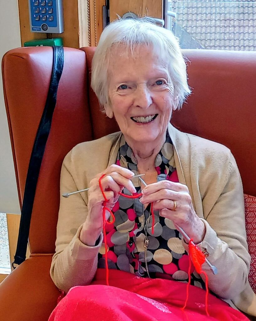 An elderly woman sits in a chair, smiling as she knits with red wool and knitting needles, making cosy gifts for Christmas Shoeboxes to share warmth with the world.