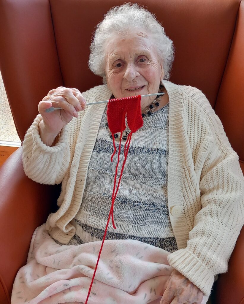 An elderly woman sits in an armchair, smiling as she knits with red wool for Wirral Christmas Shoeboxes. She is wearing a cream cardigan and has a blanket draped over her lap.