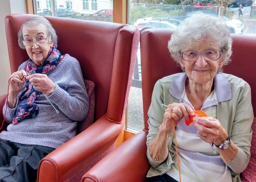 Two elderly women sit in red armchairs by a window on the Wirral, smiling and knitting colourful wool for Christmas shoeboxes.