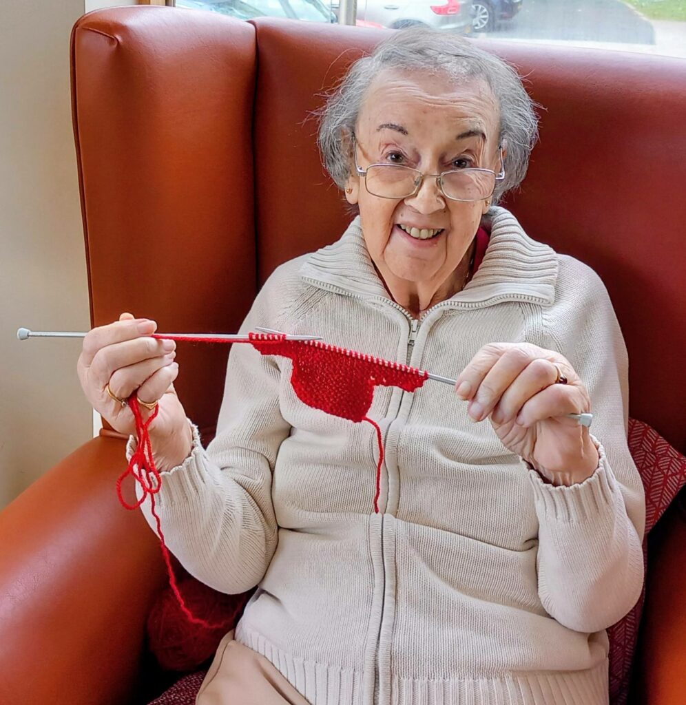 An elderly woman in Wirral sits in a chair, smiling as she holds knitting needles and works on a red item, possibly making a gift for Christmas Shoeboxes.