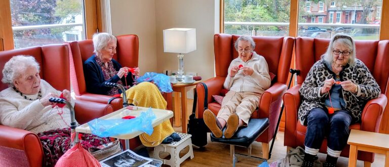 Four elderly women sit in armchairs by large windows in a cosy Wirral common room, knitting items for Christmas Shoeboxes, with yarn, mugs, and magazines on nearby tables.