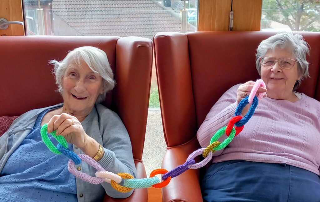 Two elderly women sit in armchairs, holding a colourful knitted chain between them, smiling at the camera in a bright Wirral home as they prepare Christmas Shoeboxes.