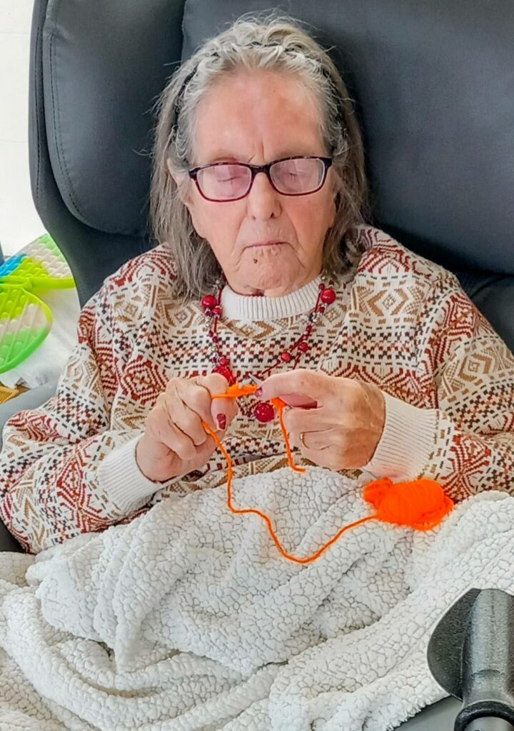 An elderly woman wearing glasses and a patterned jumper sits in a chair, knitting bright orange wool for Christmas Shoeboxes, a white blanket on her lap, in her cosy Wirral home.