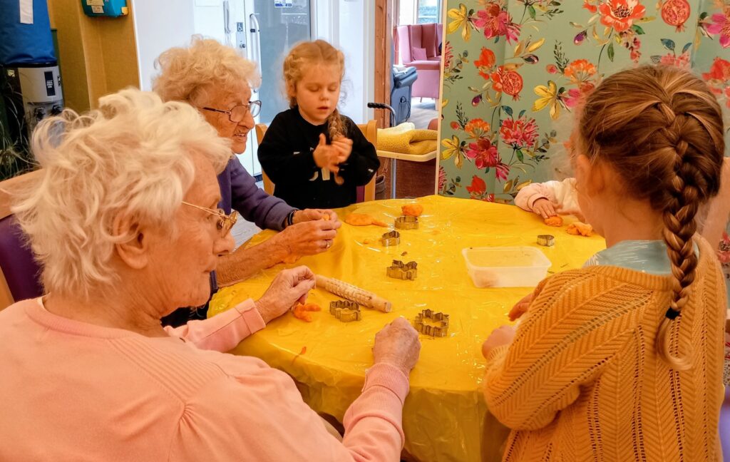 Two elderly women and three young girls sit around a yellow table, making biscuits together in a brightly decorated room, their friendship shining as brightly as the cheerful décor.