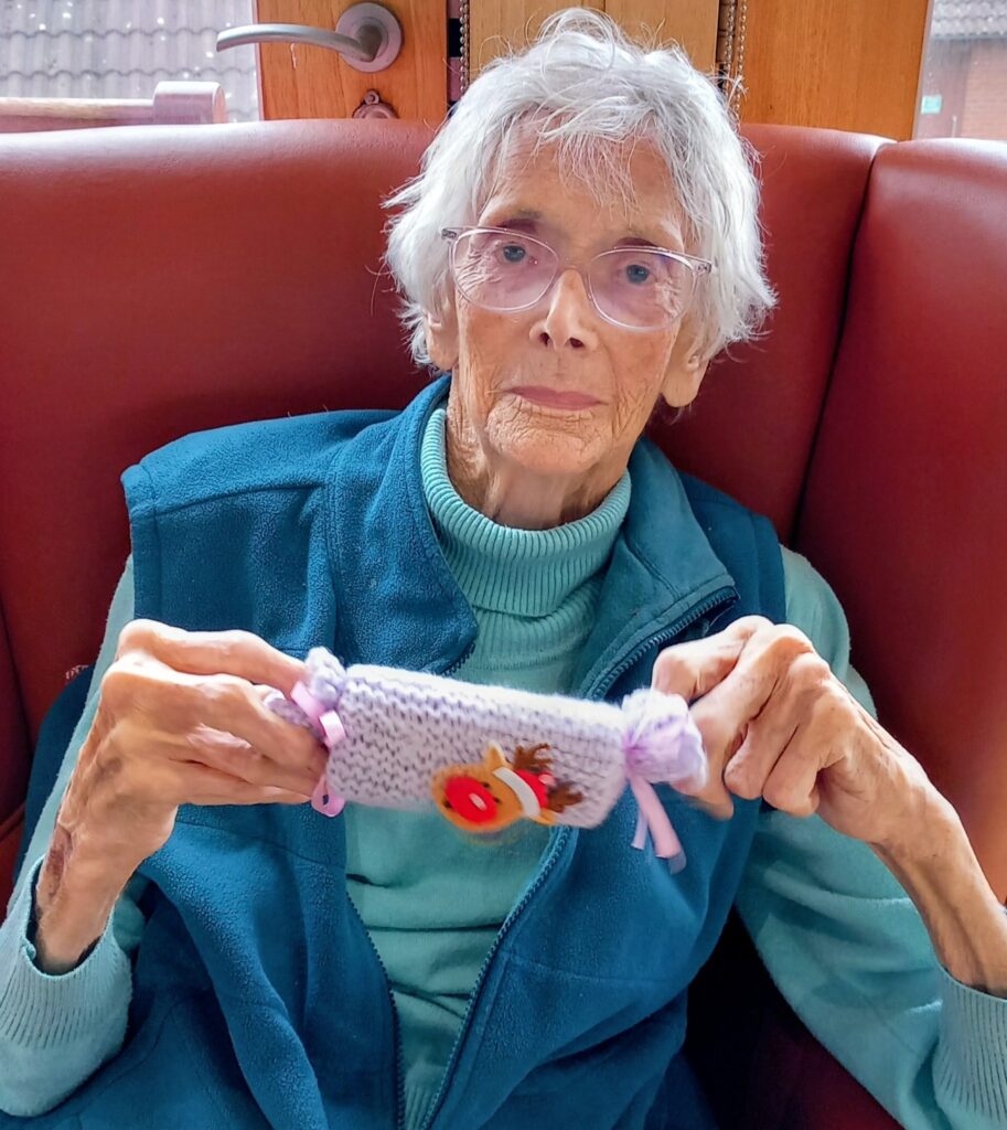Elderly woman with short white hair and spectacles sits on a red chair in Wirral, holding a small knitted item with a decorative button and ribbon, ready to place it in the Christmas shoeboxes.