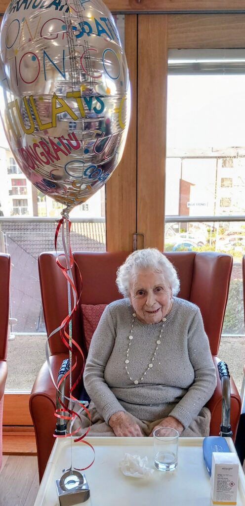 Elderly woman sits in a red chair next to a table with a drink and tissues, smiling beside a large congratulatory balloon in a bright room with big windows.