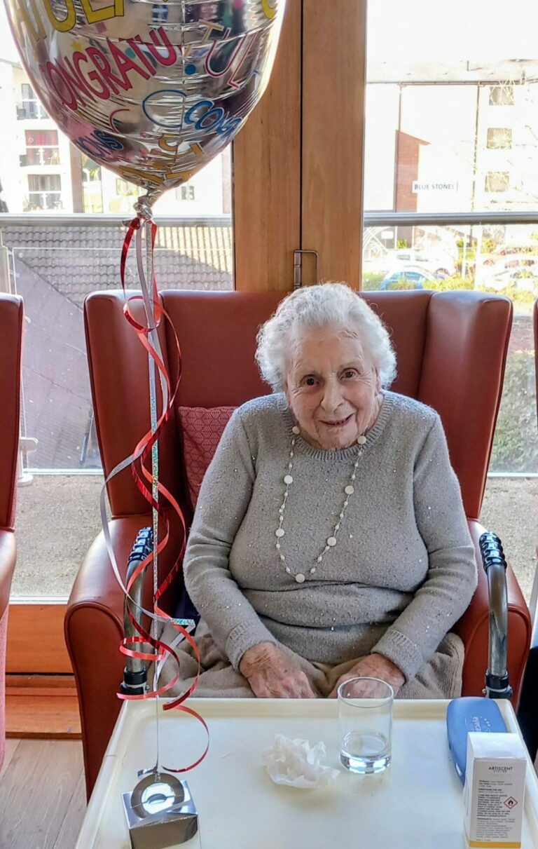 Elderly woman sits in a red chair next to a table with a drink and tissues, smiling beside a large congratulatory balloon in a bright room with big windows.